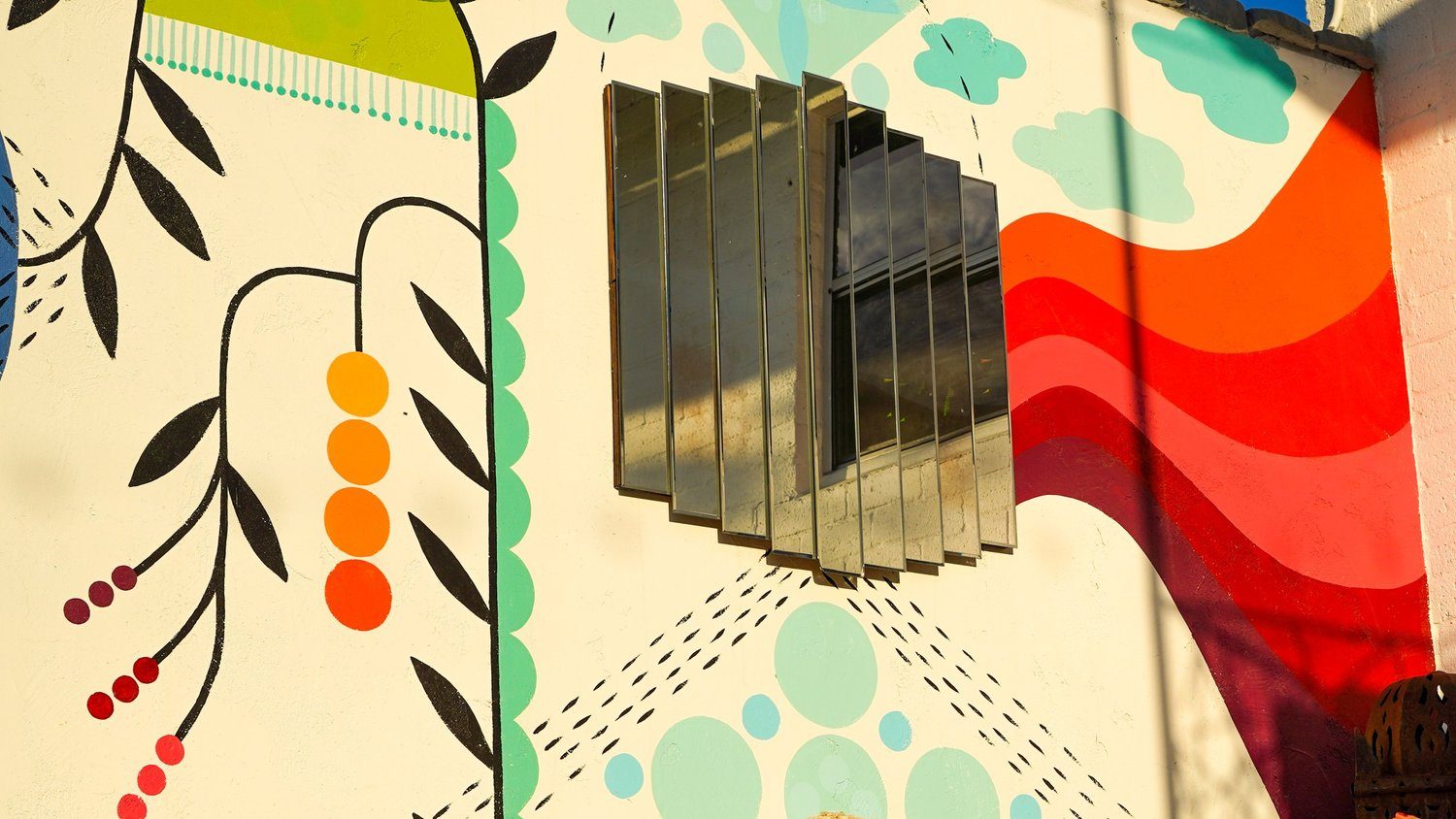 A fan of angled mirror panels framing a window on a sunlit mural wall painted with leaves, dots, and bold red waves