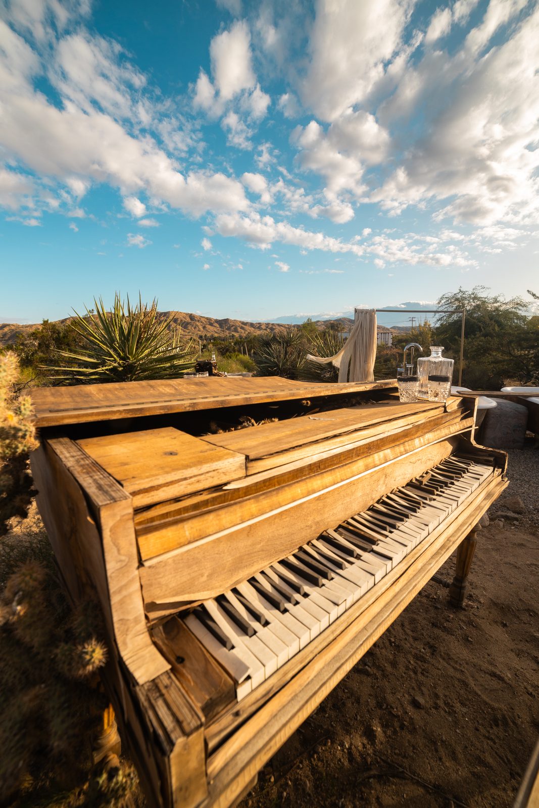 A weathered grand piano standing in the open desert beneath a dramatic sky, a crystal decanter resting on its lid